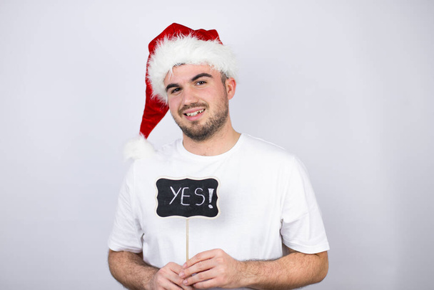 Young handsome man wearing a Santa hat over white background smiling and holding blackboard with yes word message - Photo, Image