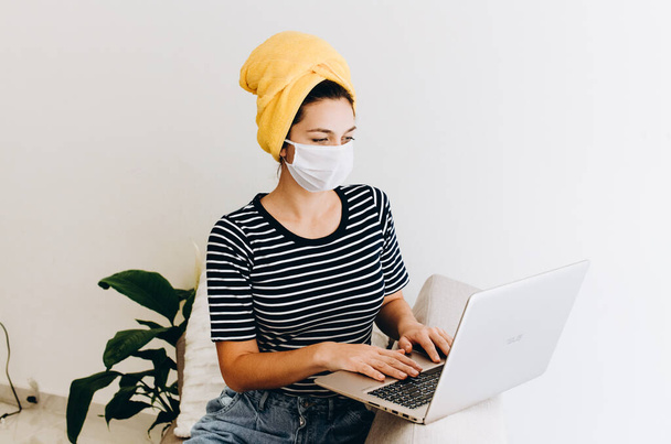Brunette Kaukasische vrouw in gele handdoek op haar hoofd werkt thuis op laptop dragen gezichtsmasker - Foto, afbeelding