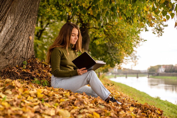 meisje zit leest een boek bij de rivier in de buurt van de boom in de herfst seizoen. - Foto, afbeelding