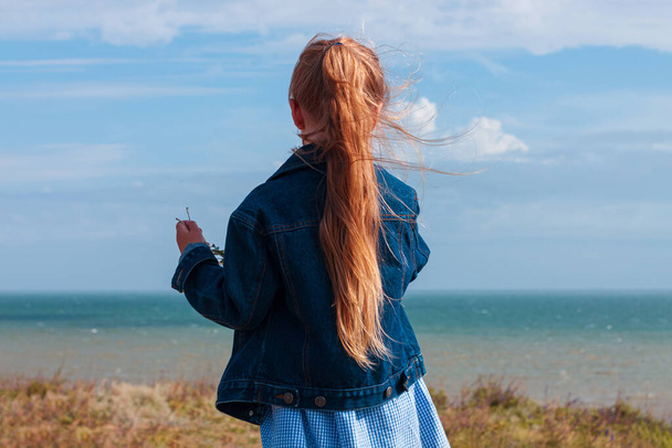 Adorable little girl in denim jacket back view on a hill with sea landscape view. Stylish child with long blonde hair on countryside cloudy sky background. Outdoor walking rural road trip faceless. - Photo, Image