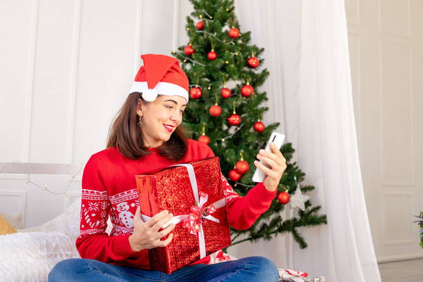 a young beautiful girl in a red sweater and a hat is making a video call with a gift on a laptop at home on a bed near a Christmas tree with a new year's interior - Photo, Image