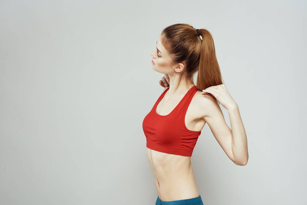 A woman in a red T-shirt on a light background is engaged in fitness gestures with her hands a slim figure  - Photo, Image