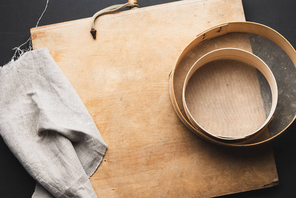 tamis rond en bois vintage, serviette en lin gris, table noire, vue de dessus, ustensiles de cuisine pour faire de la pâte - Photo, image