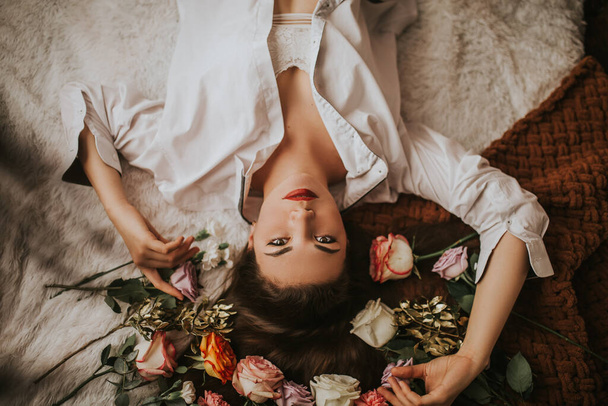 Una mujer joven yace en una cama hecha de paletas de madera con flores de diferentes tipos, fotografías en una pared de ladrillo, los rayos del sol se filtran a través de la ventana. - Foto, imagen