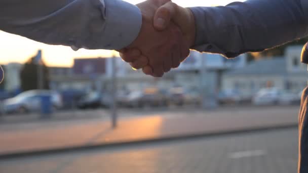 Close up of two successful businessmen greeting each other against the background of cars parking. Young colleagues meeting and shaking hands at city street on sunset. Business handshake outdoor - Footage, Video