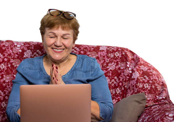 Una anciana guapa, usa un portátil y sonríe felizmente en una videollamada. Foto de una casa en un sofá con un espacio lateral vacío. - Foto, Imagen