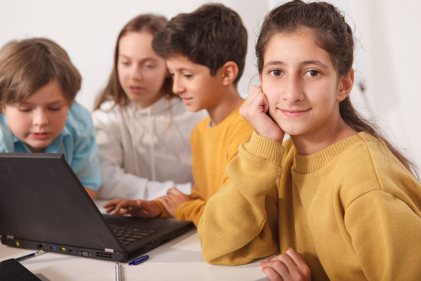 Cute Arab schoolgirl smiling to the camera, her friends using laptop at school on background - Photo, Image