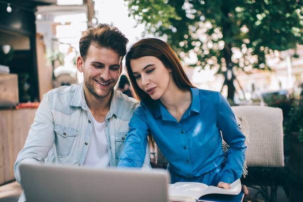 Souriant homme et femme modernes en vêtements décontractés regarder ordinateur portable et parler dans la cafétéria de la rue avec un grand arbre vert en arrière-plan - Photo, image