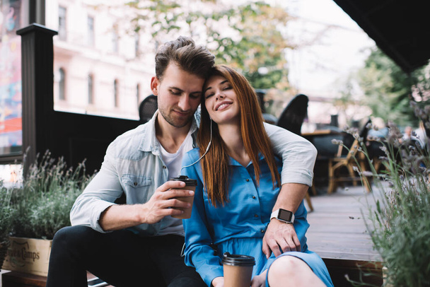 Smiling relaxed couple sitting with closed eyes on front porch drinking coffee from paper cup with man tenderly embracing woman wearing smart watch listening to music together - 写真・画像