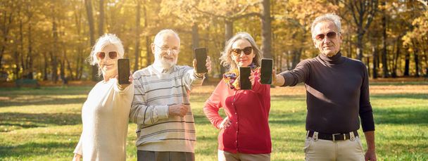 Group of senior people smiling and showing empty screens of mobile phones in autumn park - Mature friends spending time together. - Fotografie, Obrázek