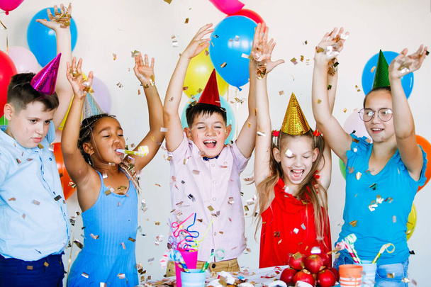Strahlende, niedliche Kinder feiern Geburtstag. Multinationale Party, Luftballons, Konfetti, Mützen, Lächeln, Teenager heben glücklich die Hände. - Foto, Bild