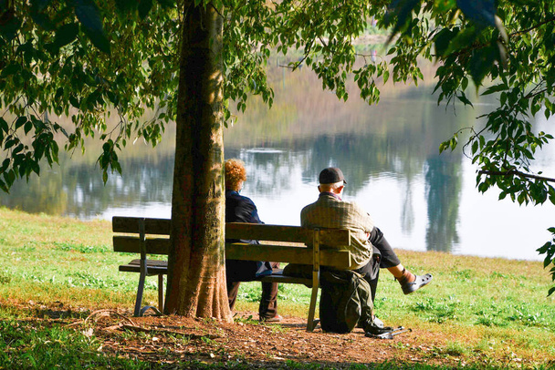 木々の紅葉を反映した水で小さな湖の近くの風景を賞賛座っている高齢者の女性と男 - 写真・画像