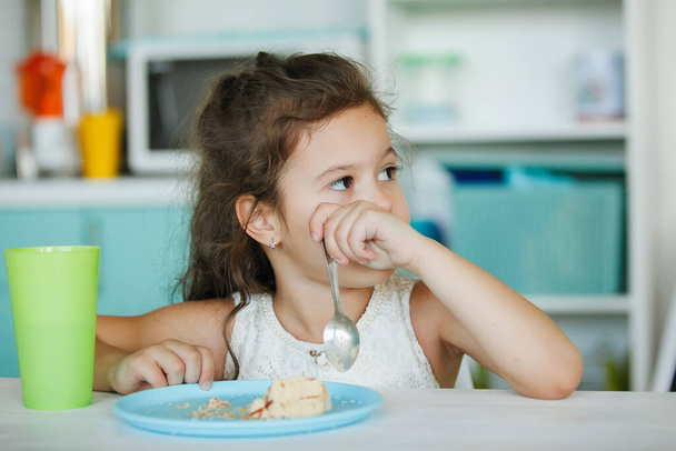 Roztomilá holka jí dort. Na stole je modrý talíř, zelená sklenice, v ruce drží lžíci. Rodinné štěstí, šťastný úsměv, dětská dovolená. - Fotografie, Obrázek