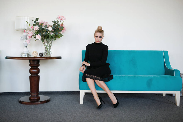Schöne Blondine sitzt auf der Couch, die Haare zum Dutt zusammengebunden, schwarzes Kleid, eleganter Stil. - Foto, Bild