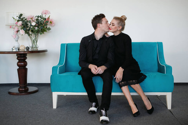 Young couple kiss on the couch. Blonde woman, red lipstick, black dress, hair in a bun. Man in a black suit and white shoes. Stylish, beautiful, daring. - Foto, Imagen