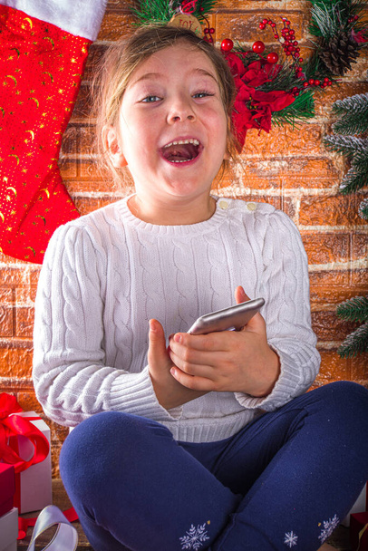 Menina bonito com Smartphone em casa com árvore de Natal, Presentes Caixas e Decoração de Natal, Covid-19 Celebrando congratulações de Natal em quarentena conceito fundo  - Foto, Imagem