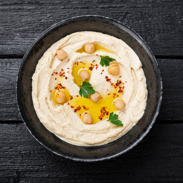 Hummus, Kichererbsen-Dip, mit Gewürzen in einem schwarzen Teller auf schwarzem Holzhintergrund. - Foto, Bild