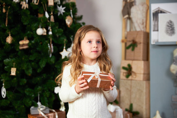 A cute little girl in a white sweater stands next to an elegant Christmas green christmas tree and holds an eco gift, looks at away and smiles - Fotoğraf, Görsel