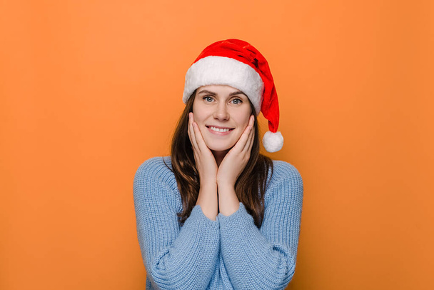 Pretty happy European woman keeps hands on cheeks, wears xmas red hat and winter sweater, poses against orange background. Cheerful lovely feminine girl feels amused. Christmas and new year concept - Фото, изображение