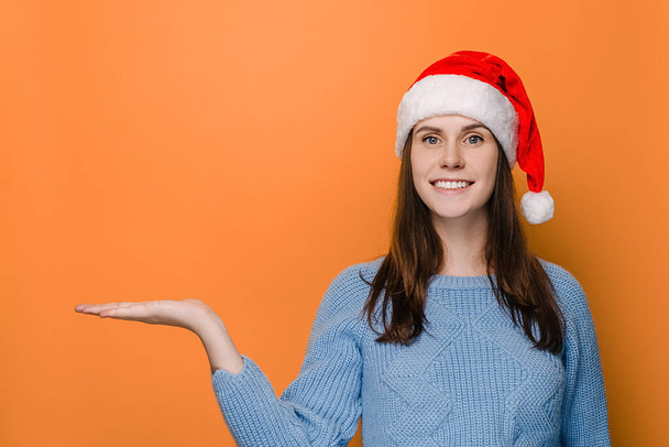 Веселая женщина с очаровательной улыбкой держит ладонь поднятой против оранжевого копирования пространства, смотрит в сторону, носит рождественскую шляпу и зимний свитер, демонстрирует что-то. С Новым годом и праздником - Фото, изображение