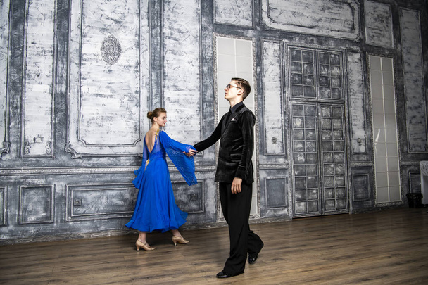 Молодой человек танцует с девушкой в синем бальном платье в сером танцевальном зале - Фото, изображение