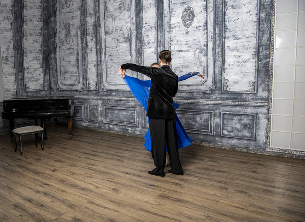 junger Mann tanzt mit einem Mädchen in einem blauen Gesellschaftskleid in einem grauen Tanzsaal - Foto, Bild
