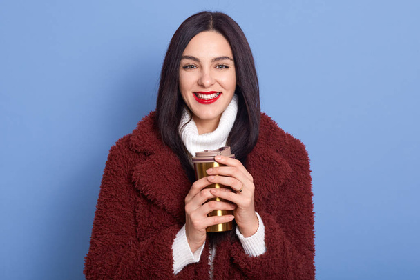 Brunette girl dresses white warm sweater and faux fur coat, posing isolated over blue background, holds coffee in thermo mug, female looks happy, looking directly at camera with cute smile. - Photo, Image