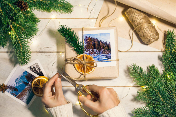 Las manos de las mujeres cortan la cuerda en regalo de Navidad en caja con fotos de montañas en estilo ecológico: papel rugoso natural, cuerda de yute, ramitas de abeto, tijeras vintage. Viajar como regalo. - Foto, imagen