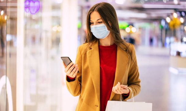 Confident trendy woman in a safety medical mask with a shopping bag and smart phone is walking in the mall during black friday - Valokuva, kuva