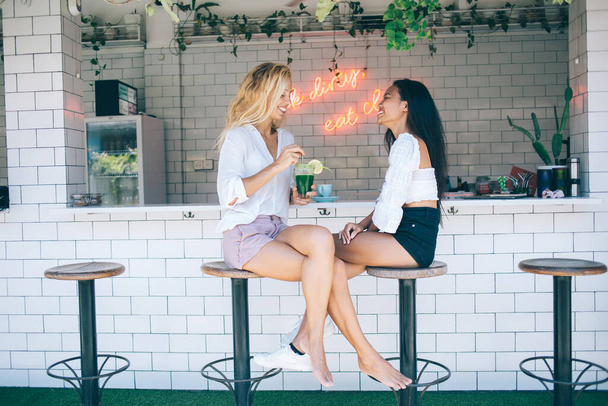 Side view of full body multiracial female friends in summer trendy outfit sitting on high chairs in cozy bar and enjoying beverages - Foto, imagen