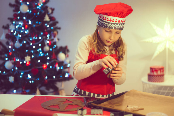 Pretty little girl decoraiting christmas cookies of different forms at home with christmas tree on background - Photo, Image