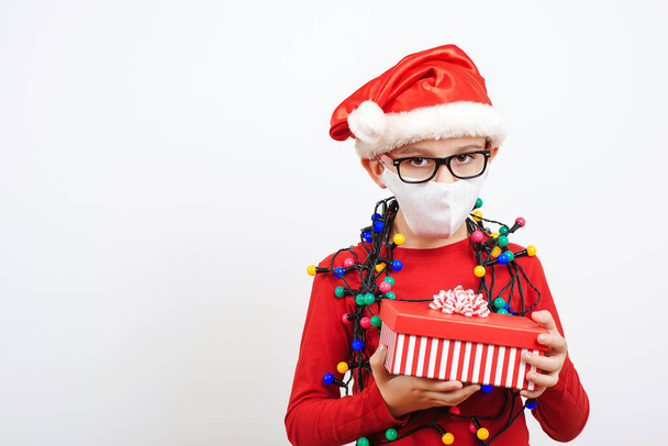 Tired little santa with christmas gifts during quarantine. Boy wearing safety face mask and santa hat. Sad child in festive garland alone with presents. Christmas during coronavirus pandemic. - Foto, afbeelding
