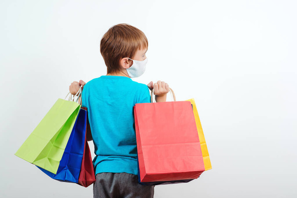 Boy in protective face mask holding shopping bags. Young shopaholic with paper bags. Seasonal sales and discounts. Kid with purchases isolated on white background. Shopping during quarantine. - Photo, Image