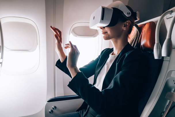 Side view of female in formal wear entertaining with virtual reality goggles and exploring air with hands while sitting in cabin of aircraft - Фото, изображение