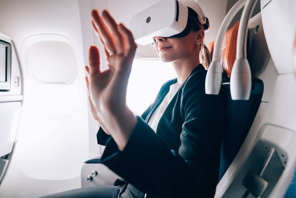 Side view of female in formal jacket using glasses of virtual reality and gesturing in air while sitting in cabin of aircraft - Fotoğraf, Görsel