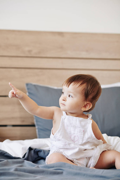Meraklı küçük çocuk yatakta oturuyor ve parmağıyla işaret ediyor. - Fotoğraf, Görsel