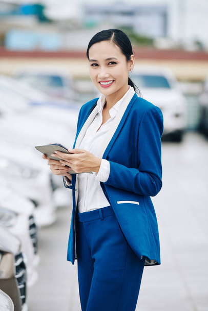 Portrait of pretty smiling young female car dealership manager working on tablet computer - Photo, Image
