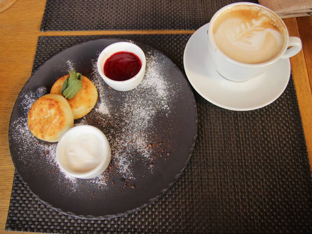 Obrázek snídaně se sýrovými palačinkami a kávou moka s krásným latte art topping.Morning jídlo podávané s cappuccino šálek v kavárně.Ruské tradiční snídaně. - Fotografie, Obrázek