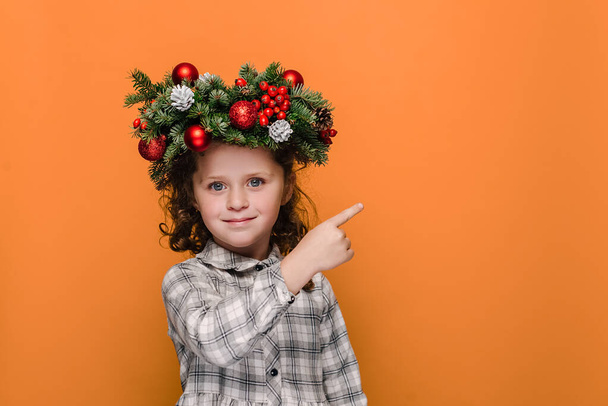 Happy little kid girl points with forefinger at free space for advertising content, wears spruce holiday wreath around head, isolated on orange studio background. New Year and Christmas concept - Foto, immagini