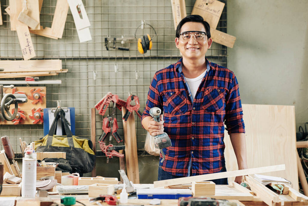 Portrét šťastného zralého asijského tesaře s elektrickou vrtačkou stojící na pracovním stole pokrytém dřevěnými polotovary a nástroji - Fotografie, Obrázek