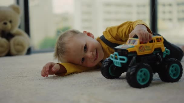 Niño alegre acostado en la alfombra con camión de juguete. Bebé jugando con coche de juguete en casa. - Imágenes, Vídeo