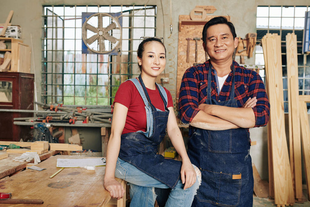 Portrait of happy Vietnamese carpenters in denim aprons standing in workshop - Photo, Image