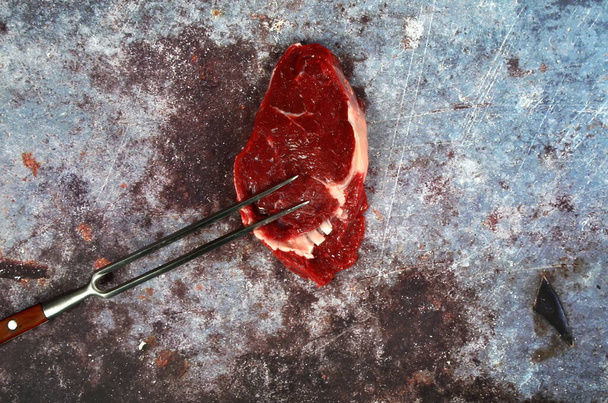 Ωμό κρέας με ψήσιμο μαχαίρι και πιρούνι, μπριζόλα βοείου κρέατος σε σκούρο ρουστίκ φόντο, κορυφαία άποψη, αντίγραφο χώρου - Φωτογραφία, εικόνα