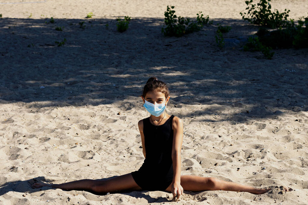 A gymnast girl sports in a protective mask, against the backdrop of the sea and the beach. Performs a stretch exercise. Fitness. The concept of safe outdoor training in a medical mask and in sportswear. Quarantine. - Photo, Image