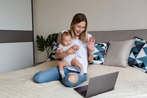 Telecommuting in quarantine. Mother showing baby to friends at the video chat. Stylish young freelancer working from home. Social distancing during coronavirus covid-19 pandemic concept. - Zdjęcie, obraz