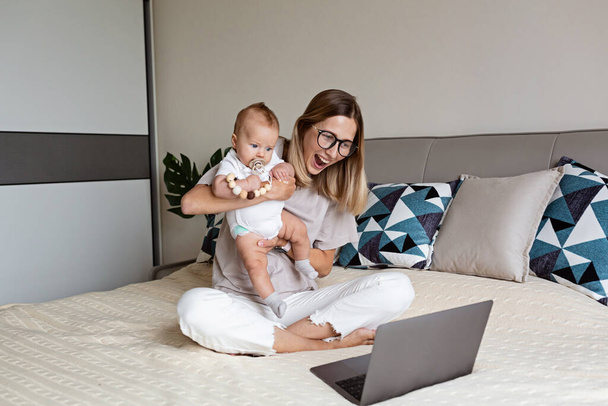 Telecommuting in quarantine. Mother showing baby to friends at the video chat. Stylish young freelancer working from home. Social distancing during coronavirus covid-19 pandemic concept. - Photo, Image