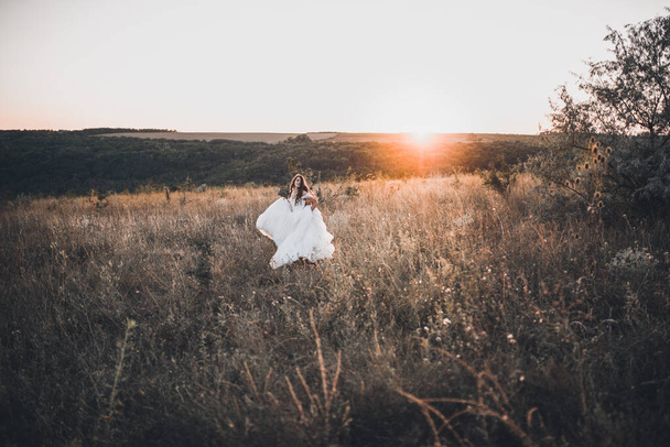 Bride in wedding white dress walking on meadow in summer at sunset - Фото, изображение