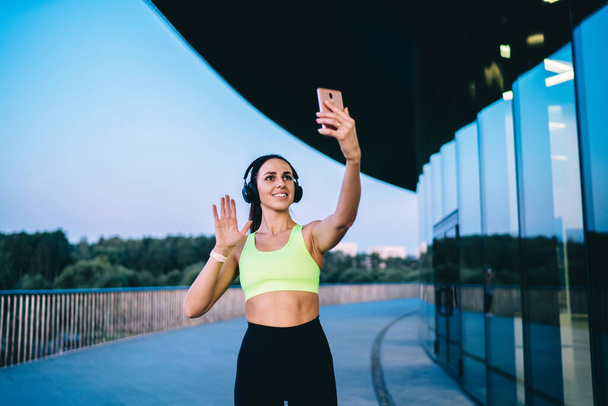 Content slim female athlete wearing trendy bra and leggings recording video on smartphone and smiling while standing on city street during training and listening to music in headphones - Zdjęcie, obraz