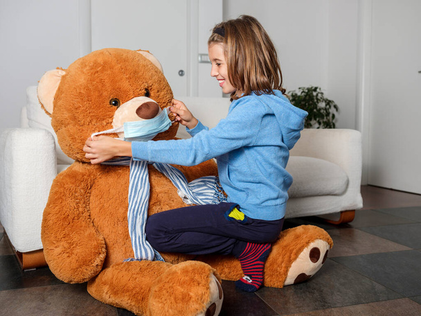 Child in lockdown makes his teddy bear wear the coronavirus mask. Child in home quarantine playing with his big teddy bear wearing a medical mask against viruses during coronavirus outbreak. Flu, illness, pandemic concept. - Photo, Image