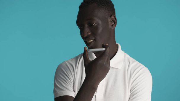 Молодой привлекательный афроамериканец записывает голосовое сообщение другу с помощью смартфона на цветном фоне - Фото, изображение